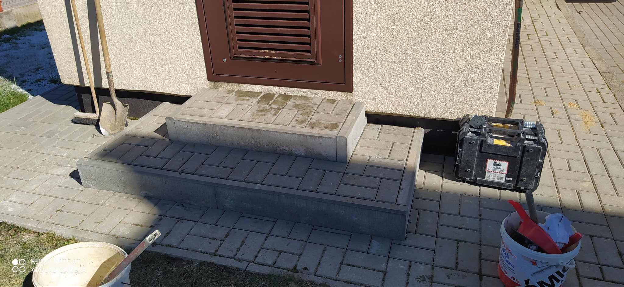 Ułożone schody wejściowe z kostki brukowej w odcieniach szarości, narzędzia pracy obok, na zewnątrz budynku z beżową elewacją i brązową kratką wentylacyjną.