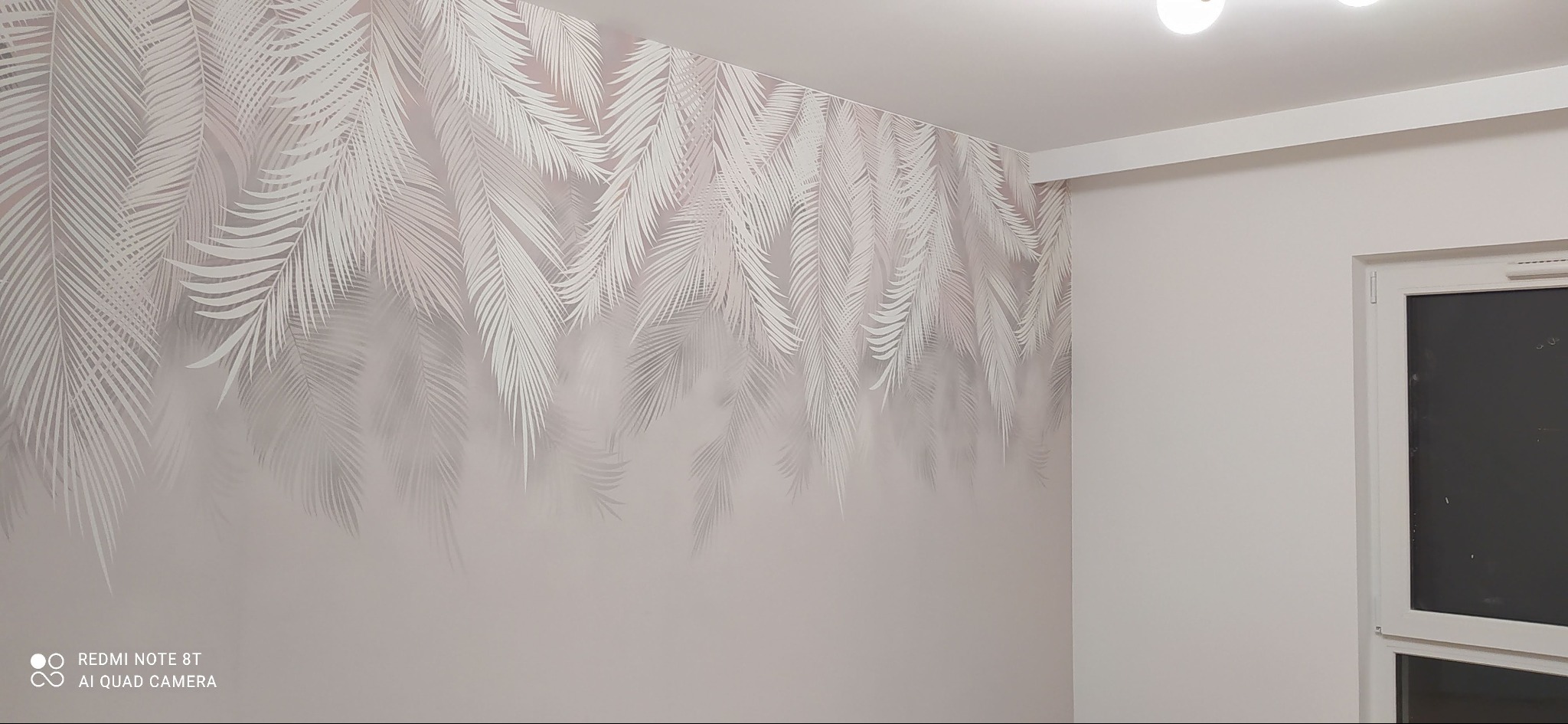 Ściana z motywem liści palmowych w odcieniach szarości, oświetlenie sufitowe i okno w rogu pokoju. Minimalistyczny design wnętrza.