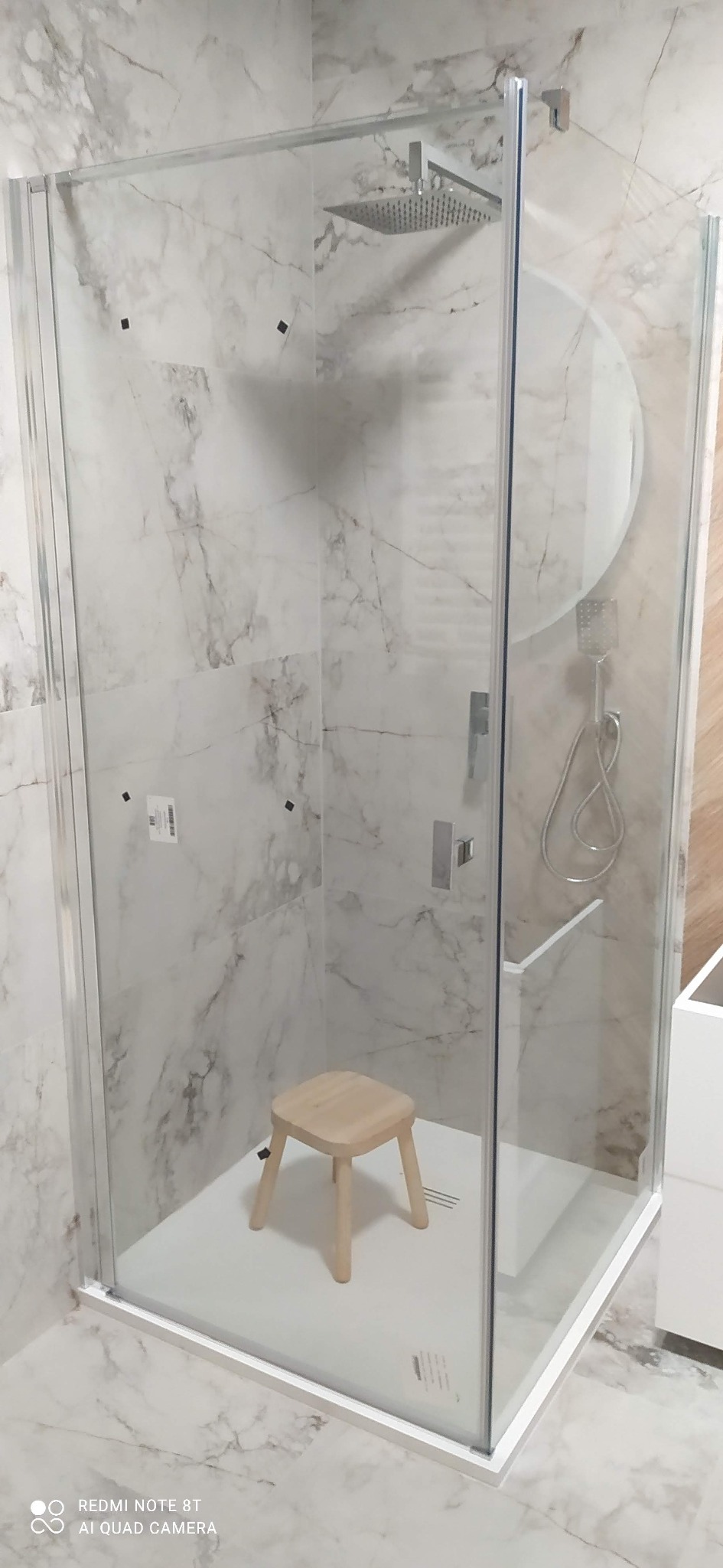 Nowoczesna kabina prysznicowa z marmurowymi płytkami, deszczownicą i drewnianym stołkiem. Minimalistyczny design, szklane ściany, jasne wnętrze.