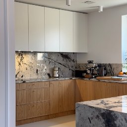 BATTEMEBLE DARIUSZ BATTE - Jasna kuchnia z drewnianymi szafkami dolnymi i białymi górnymi, blat i ściana z marmuru. Na blacie ekspres do kawy i czajnik. Wyspa kuchenna z marmurowym blatem.