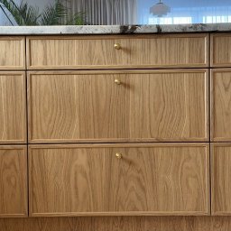 BATTEMEBLE DARIUSZ BATTE - Fronty szafek kuchennych z jasnego drewna z subtelnymi, złotymi gałkami. Blat z jasnego kamienia, w tle widoczne rośliny i lampy.