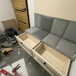 BATTEMEBLE DARIUSZ BATTE - Siedzisko z szufladami w trakcie montażu. Widoczne narzędzia, odkurzacz przemysłowy i niedokończone wykończenie podłogi. Szare panele siedziska i kremowe fronty szuflad.