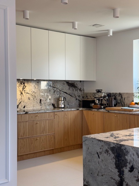 Jasna kuchnia z drewnianymi szafkami dolnymi i białymi górnymi, blat i ściana z marmuru. Na blacie ekspres do kawy i czajnik. Wyspa kuchenna z marmurowym blatem.