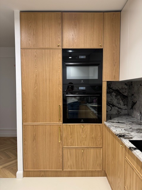 Drewniana zabudowa kuchenna z piekarnikiem i mikrofalówką w Warka. Jasne fronty z subtelnymi, złotymi uchwytami. Marmurowy blat dodaje elegancji.