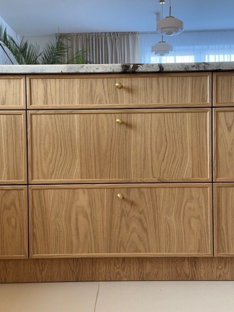 Fronty szafek kuchennych z jasnego drewna z subtelnymi, złotymi gałkami. Blat z jasnego kamienia, w tle widoczne rośliny i lampy.