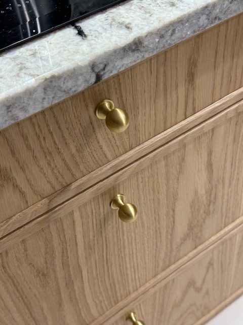 Jasne drewniane fronty szuflad z gałkami w kolorze złotym, widoczne pod blatem z jasnego marmuru. Detal wykończenia mebli kuchennych na wymiar.