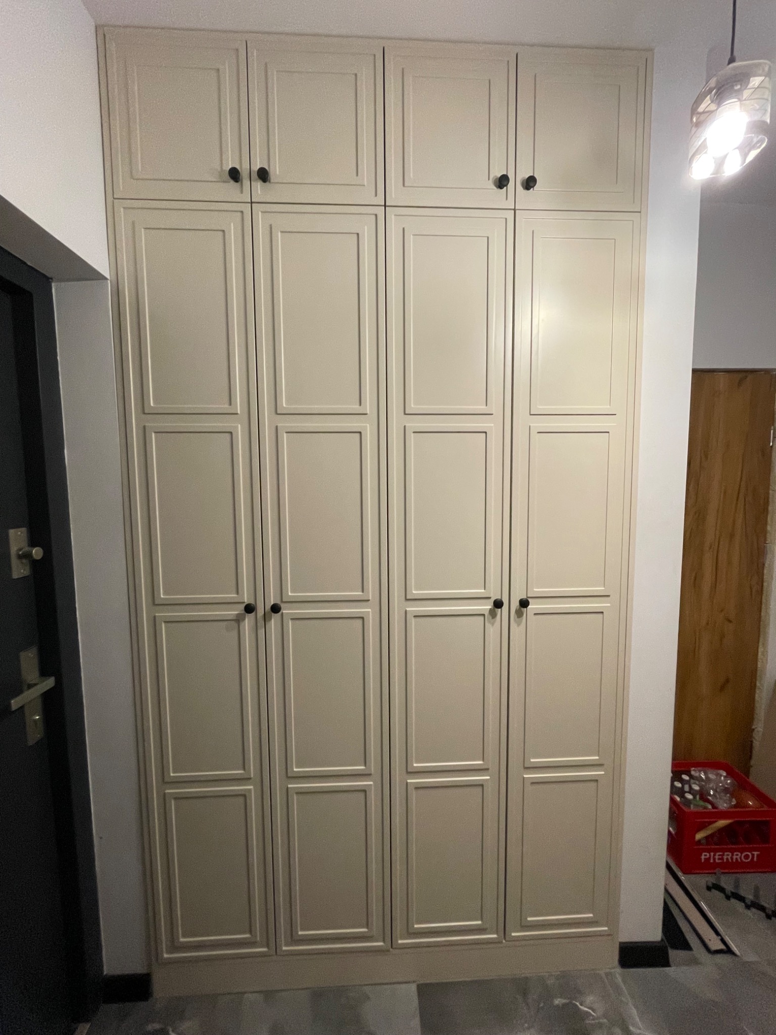 Czterodrzwiowa, beżowa szafa z czarnymi gałkami, z panelami dekoracyjnymi na drzwiach, ustawiona w korytarzu przy drzwiach wejściowych. Widoczna lampa sufitowa.