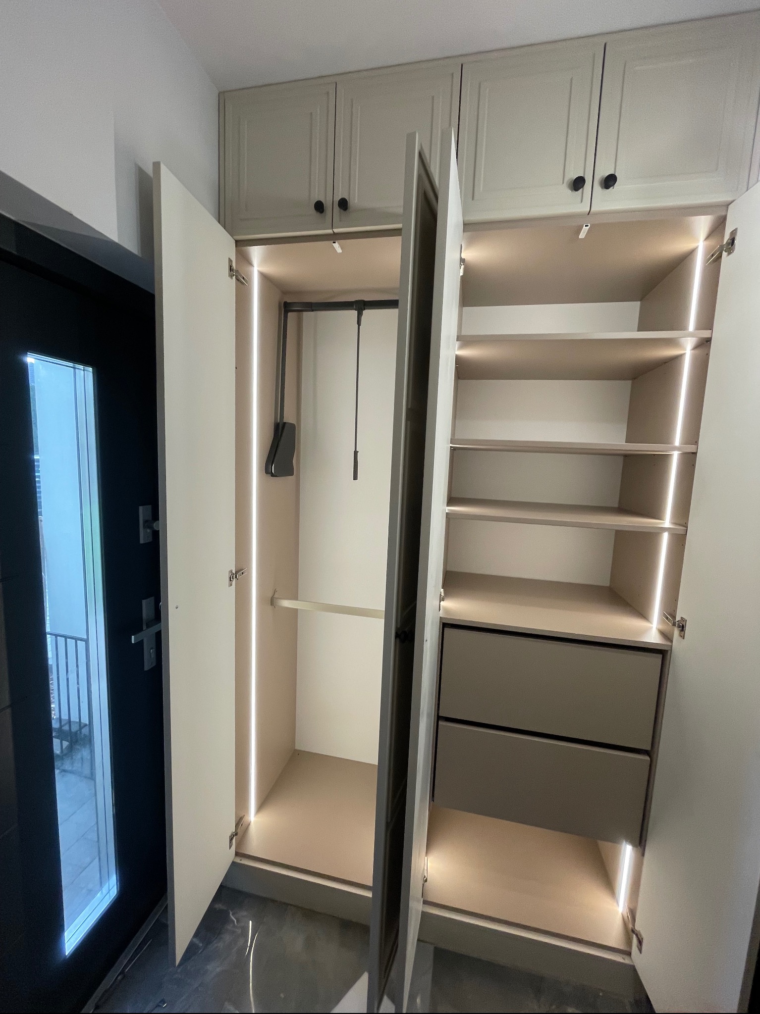 Jasna szafa wnękowa z oświetleniem LED, otwarte drzwi ukazują półki i szuflady. Widoczny system wieszaków. Obok drzwi wejściowe z widokiem na balkon.