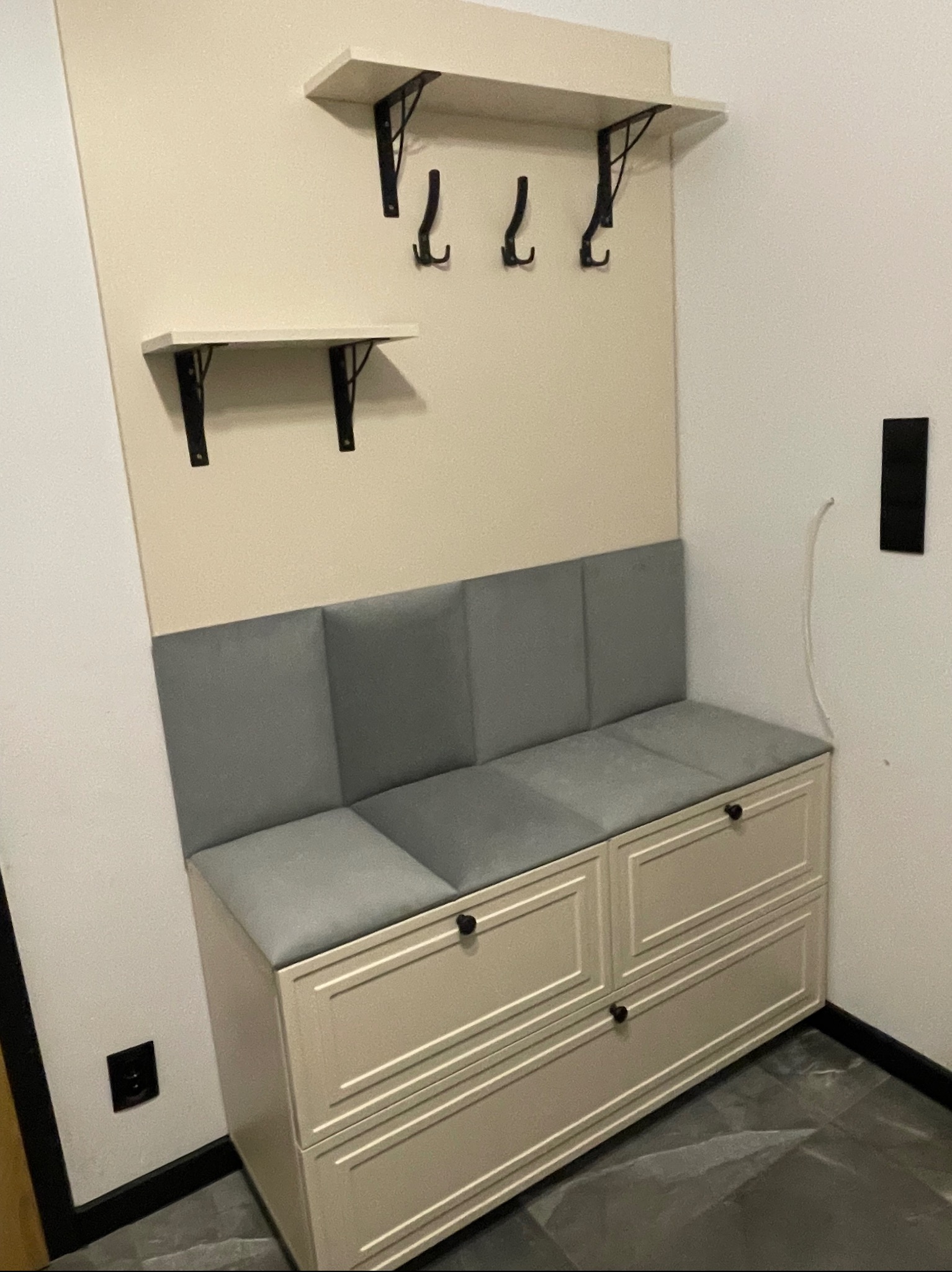 Funkcjonalna zabudowa przedpokoju w Warka: siedzisko z szufladami, wieszaki i półki na ścianie w kremowej kolorystyce. Nowoczesny design, szare poduszki siedziska.