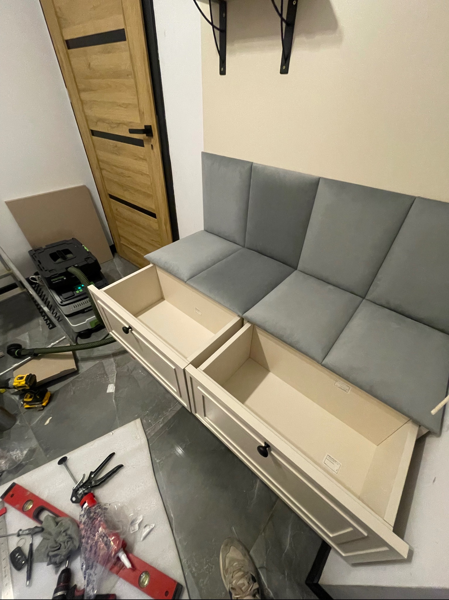 Siedzisko z szufladami w trakcie montażu. Widoczne narzędzia, odkurzacz przemysłowy i niedokończone wykończenie podłogi. Szare panele siedziska i kremowe fronty szuflad.