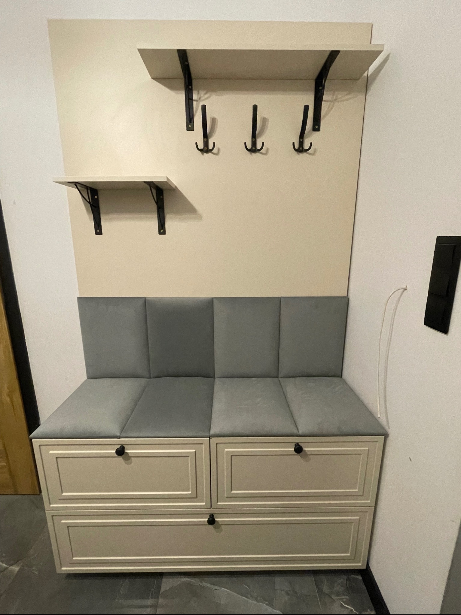 Funkcjonalna zabudowa przedpokoju: siedzisko z szufladami, wieszaki na ścianie i półka. Jasna kolorystyka, minimalistyczny design, czarne akcenty.