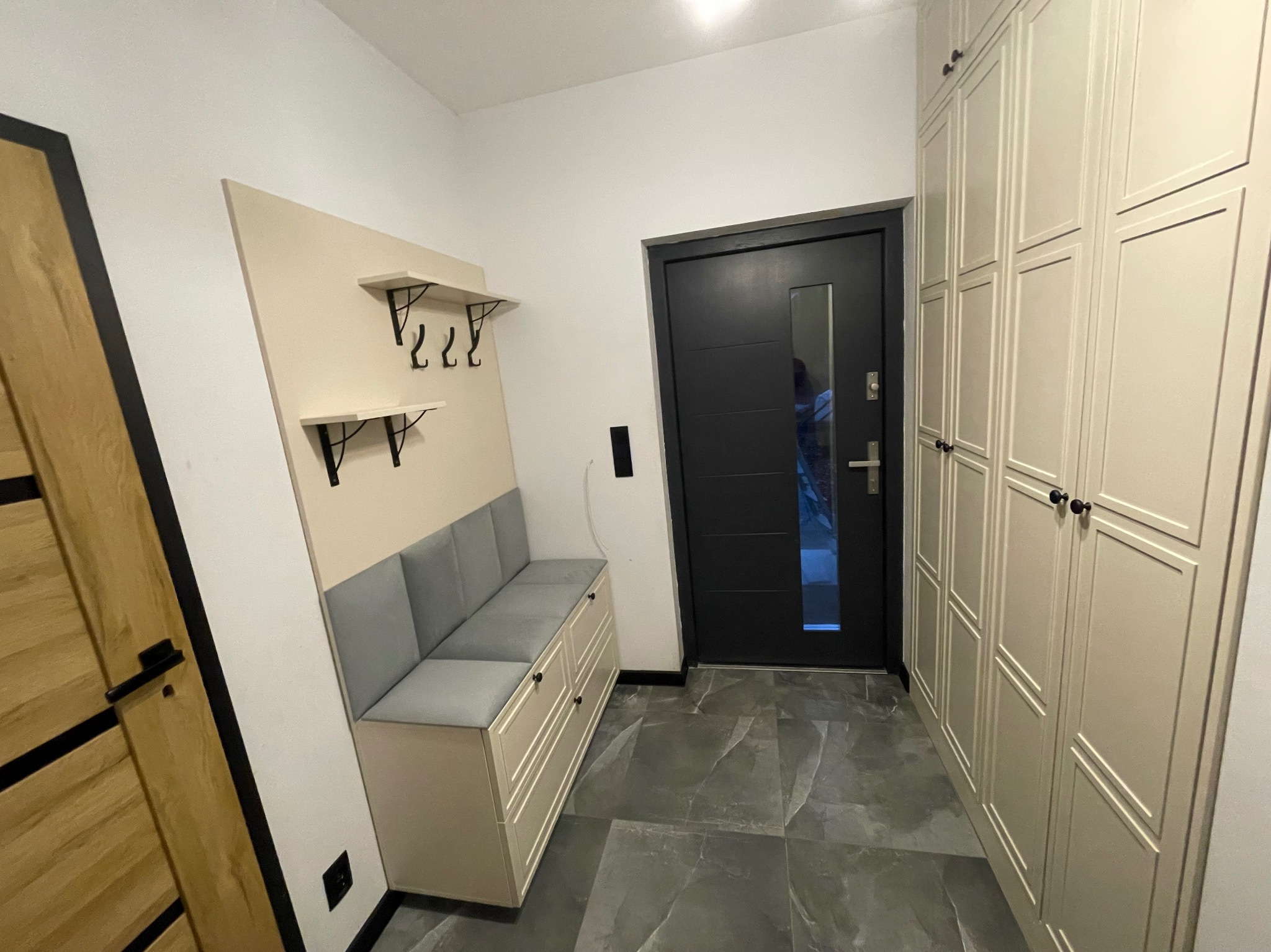 Jasny przedpokój z zabudową: szafa z frezowanymi frontami, siedzisko z wieszakami na ścianie. Szare płytki podłogowe i czarne akcenty wykończeniowe.