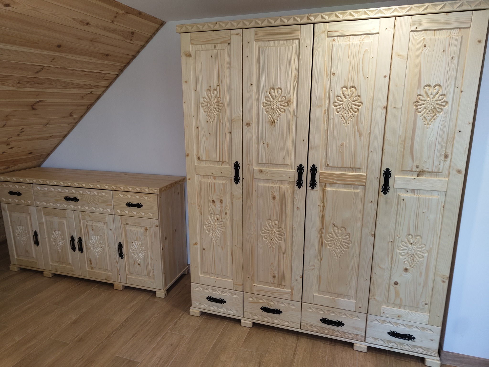 Drewniany komplet mebli z motywami góralskimi: szafa trzydrzwiowa i komoda, w jasnym odcieniu drewna, na tle poddasza z drewnianą boazerią.