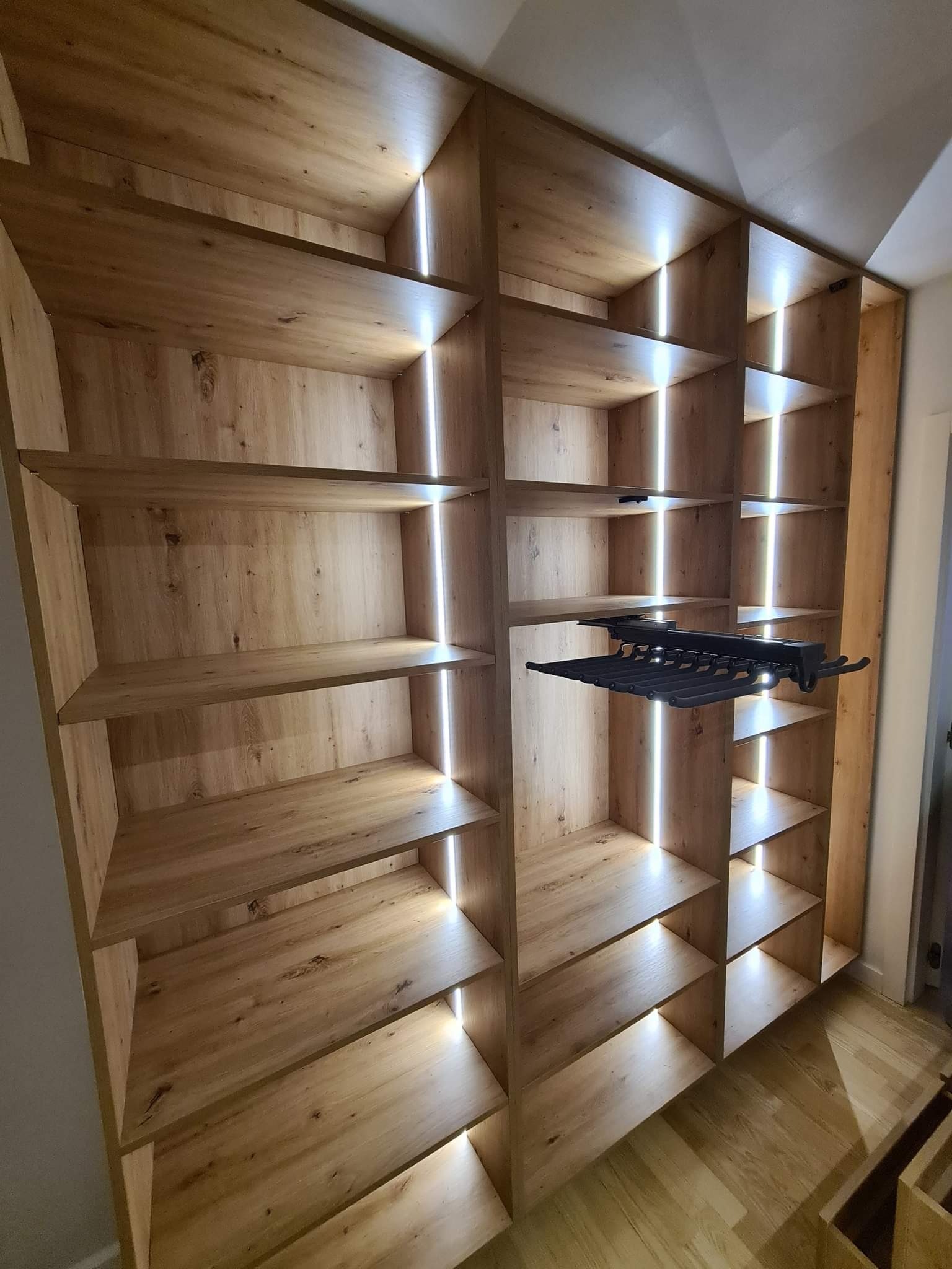 Drewniany regał z oświetleniem LED, widoczny wysuwany wieszak na ubrania. Naturalne wykończenie drewna, ciepłe światło. Nowoczesny design.