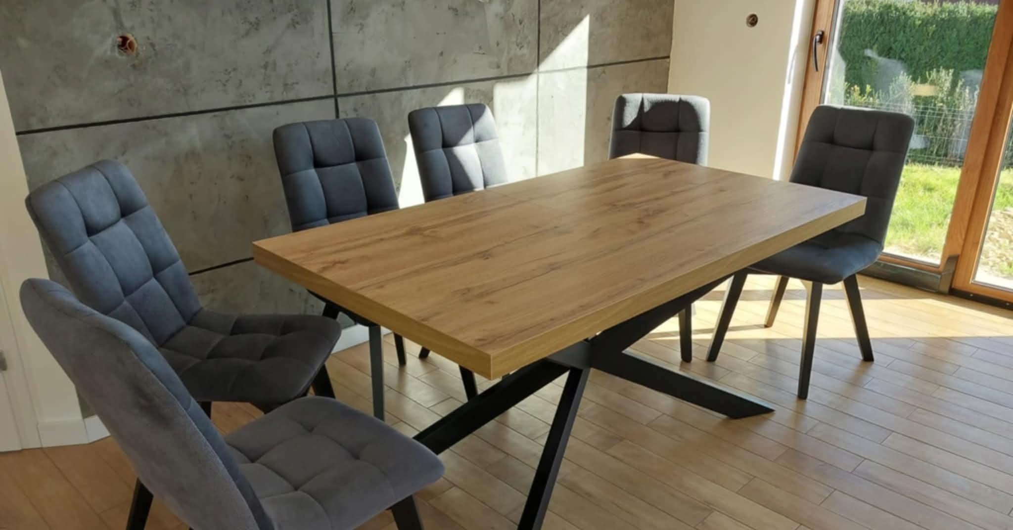 Stół z drewnianym blatem i czarnymi nogami w kształcie litery X, otoczony szarymi krzesłami na tle betonowej ściany i okna z widokiem na zieleń.
