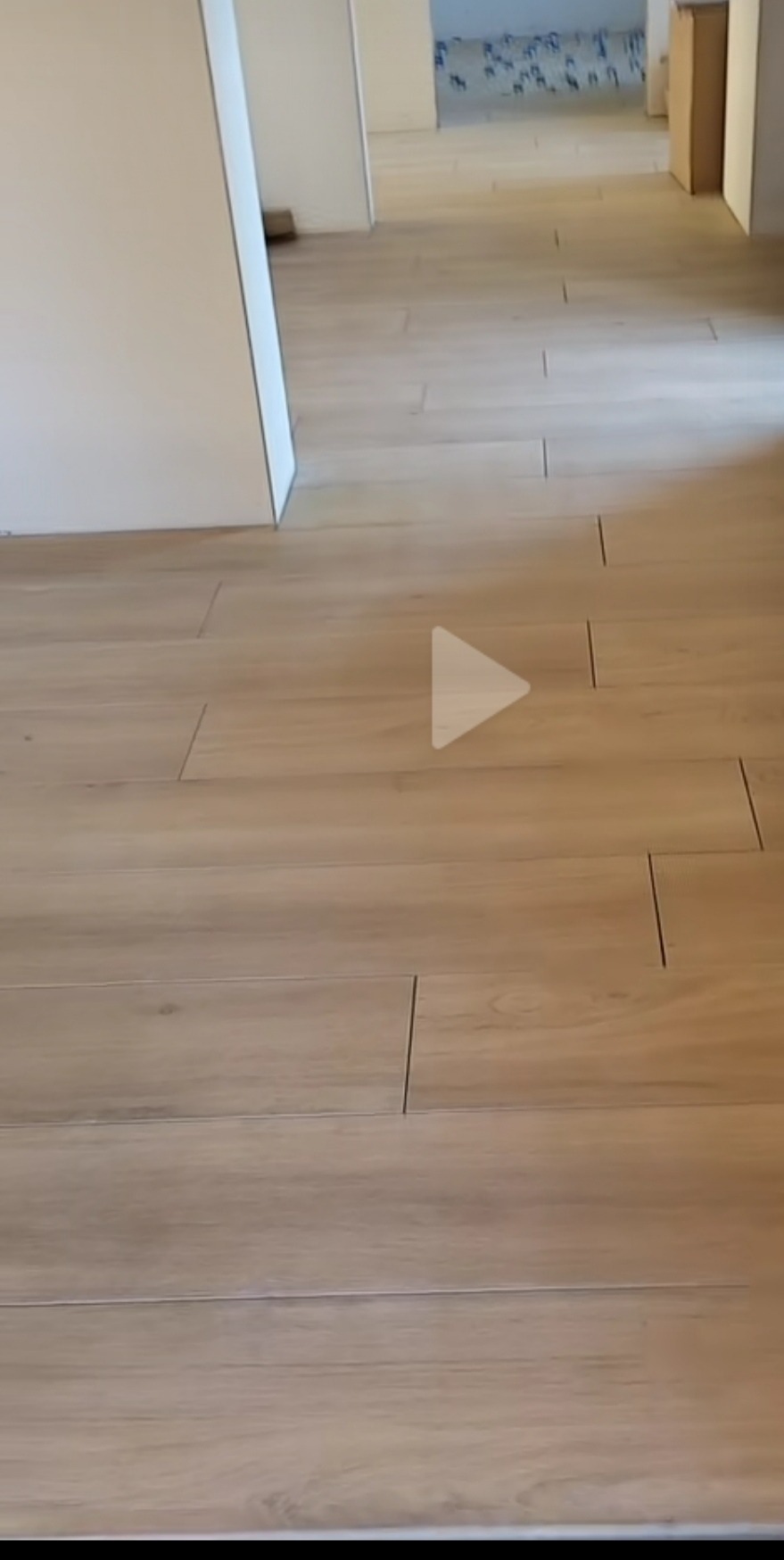 Beżowe, podłużne płytki podłogowe ułożone w jasnym wnętrzu, widoczne fugi. W tle pomieszczenie z przygotowaną podłogą. Minimalistyczny, nowoczesny styl.