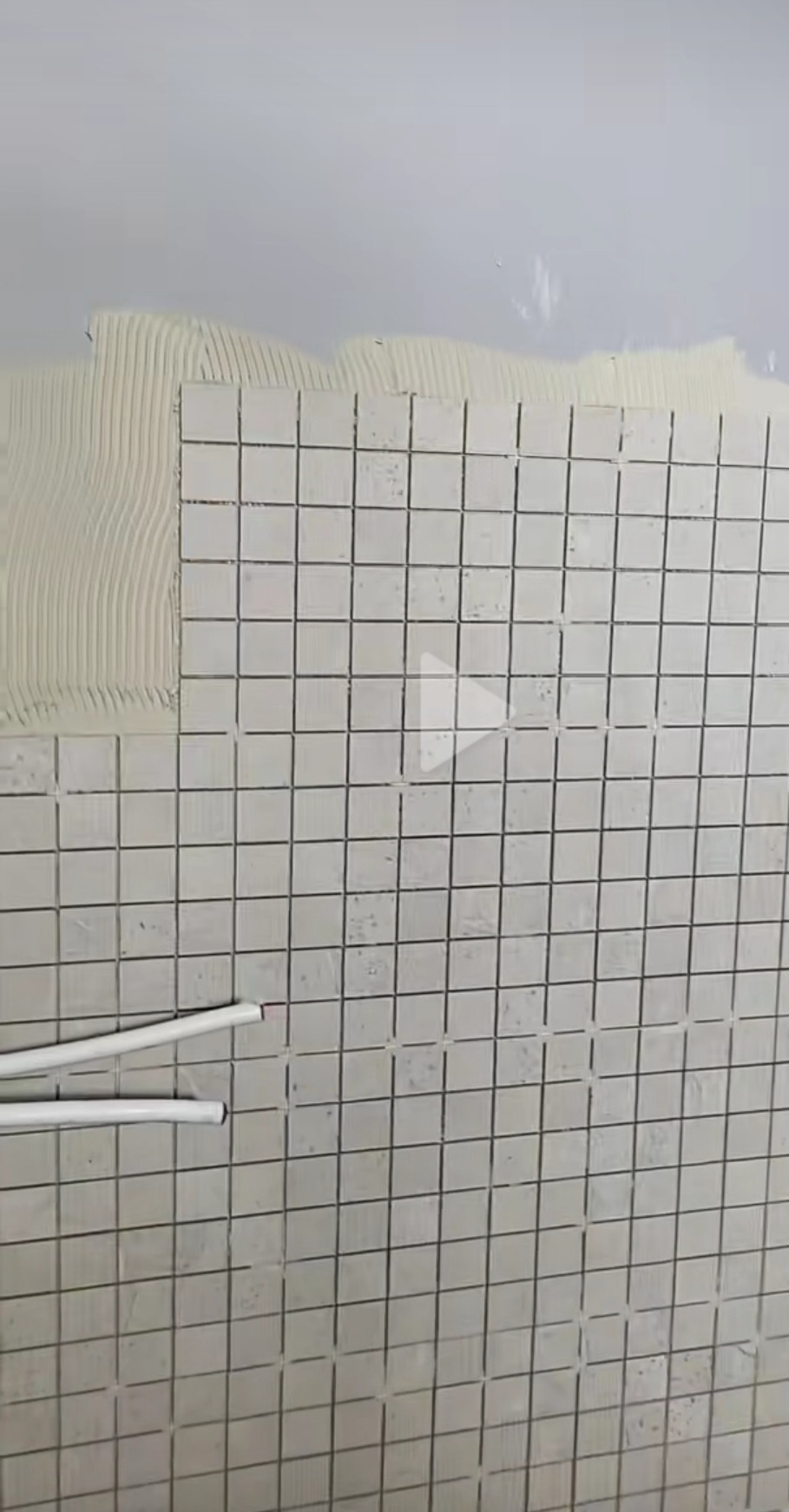 Ściana w trakcie wykańczania mozaiką ceramiczną, widoczne rury instalacyjne i nałożony klej. Prace wykończeniowe w łazience.