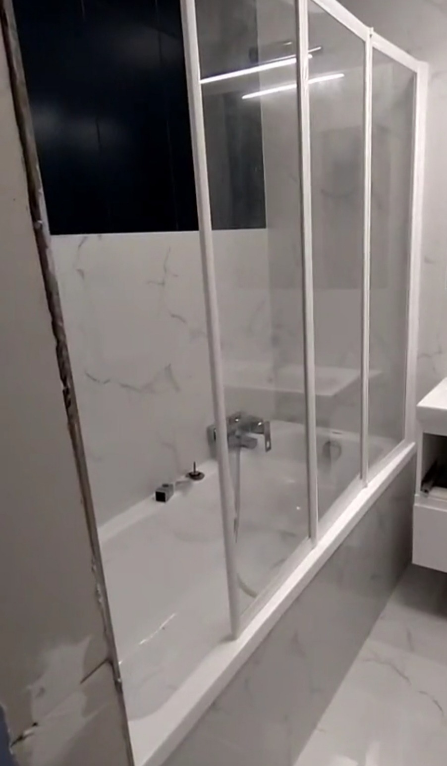 Wykończona łazienka: biała wanna z parawanem nawannowym, marmurowe płytki i czarny panel ścienny. Widoczne elementy armatury i niedokończone wykończenie ściany.