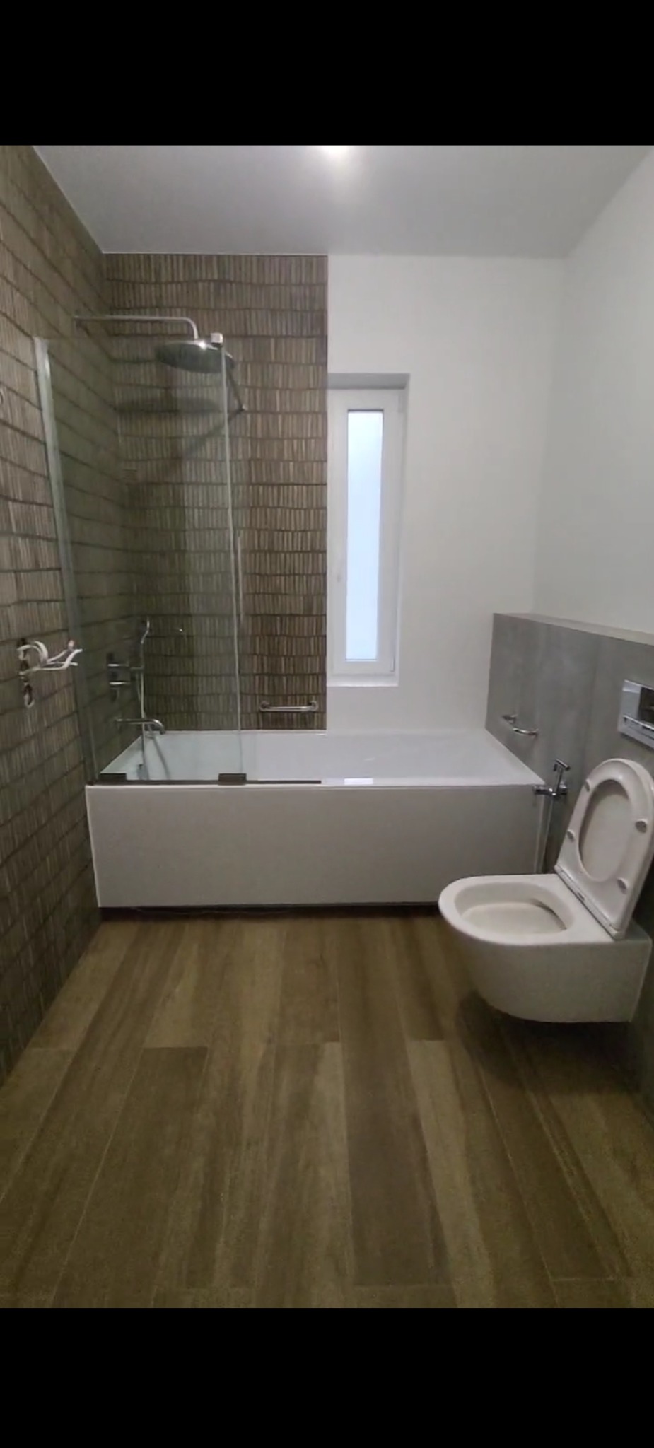 Nowoczesna łazienka po remoncie: brązowe płytki imitujące drewno, biała wanna z parawanem i podwieszana toaleta. Minimalistyczny design, stonowana kolorystyka.