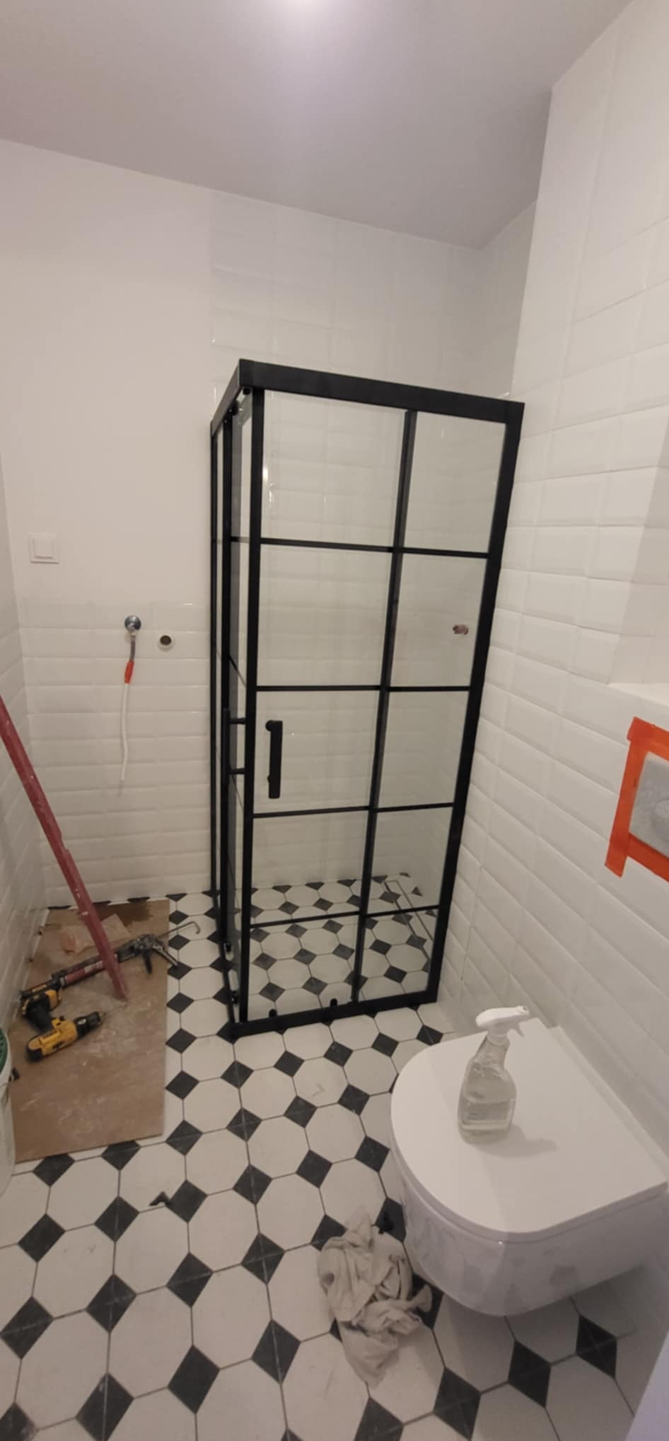 Kabina prysznicowa z czarnymi profilami na tle białych płytek i podłogi w romby. Widoczne narzędzia i akcesoria montażowe.