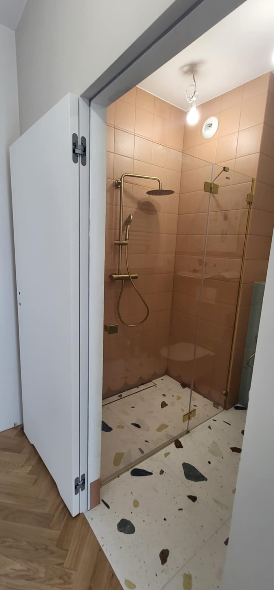 Wykończona łazienka z kabiną prysznicową typu walk-in, beżowymi płytkami i podłogą w stylu lastryko. Widoczny złoty zestaw natryskowy i biała szafka.