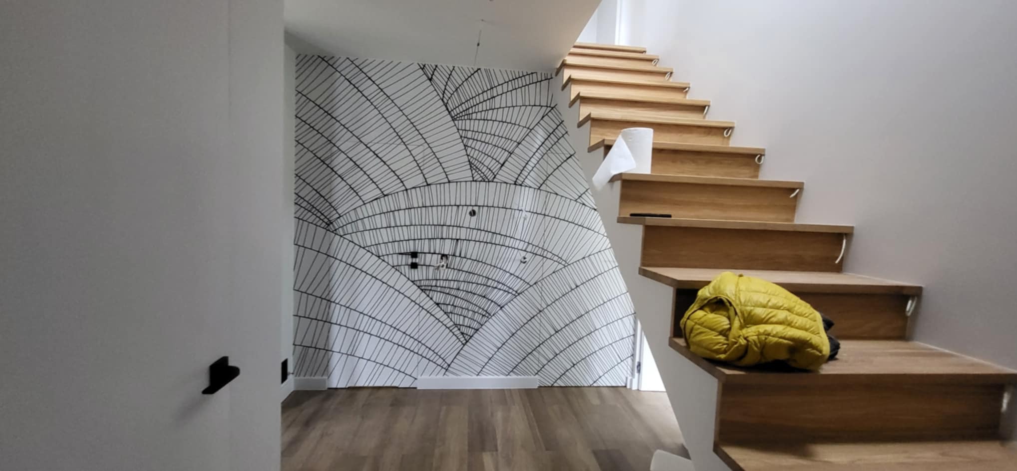 Nowoczesne schody drewniane z żółtą kurtką, ściana z geometryczną tapetą w odcieniach bieli i czerni. Widoczne gniazdka elektryczne na ścianie.