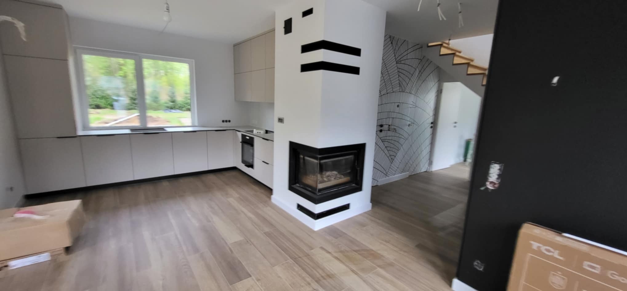 Nowoczesne wnętrze: jasna kuchnia z drewnianą podłogą, kominkiem z czarnymi akcentami i schodami w tle. Minimalistyczny design.