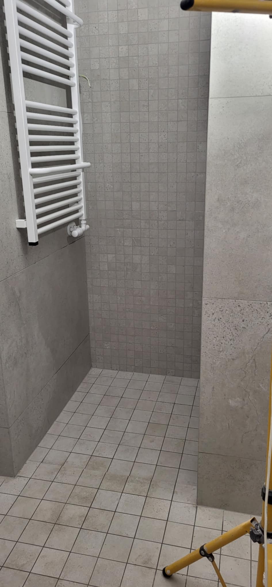 Wykończona łazienka z grzejnikiem drabinkowym i mozaiką w odcieniach szarości. Widoczny fragment drabiny malarskiej.