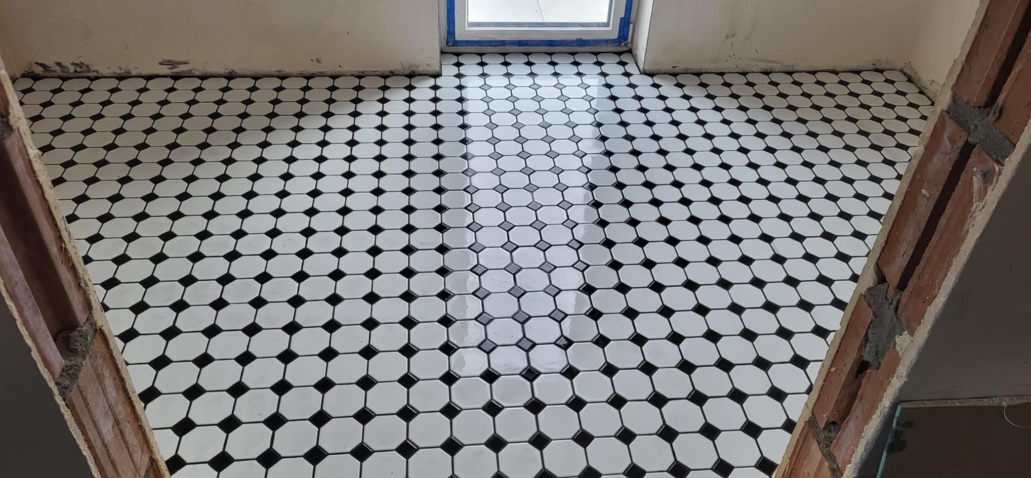 Ułożona podłoga z białych, ośmiokątnych płytek z czarnymi kwadratami w narożach, widok z góry, surowy stan wykończenia ścian, ceglany fragment ściany po prawej.