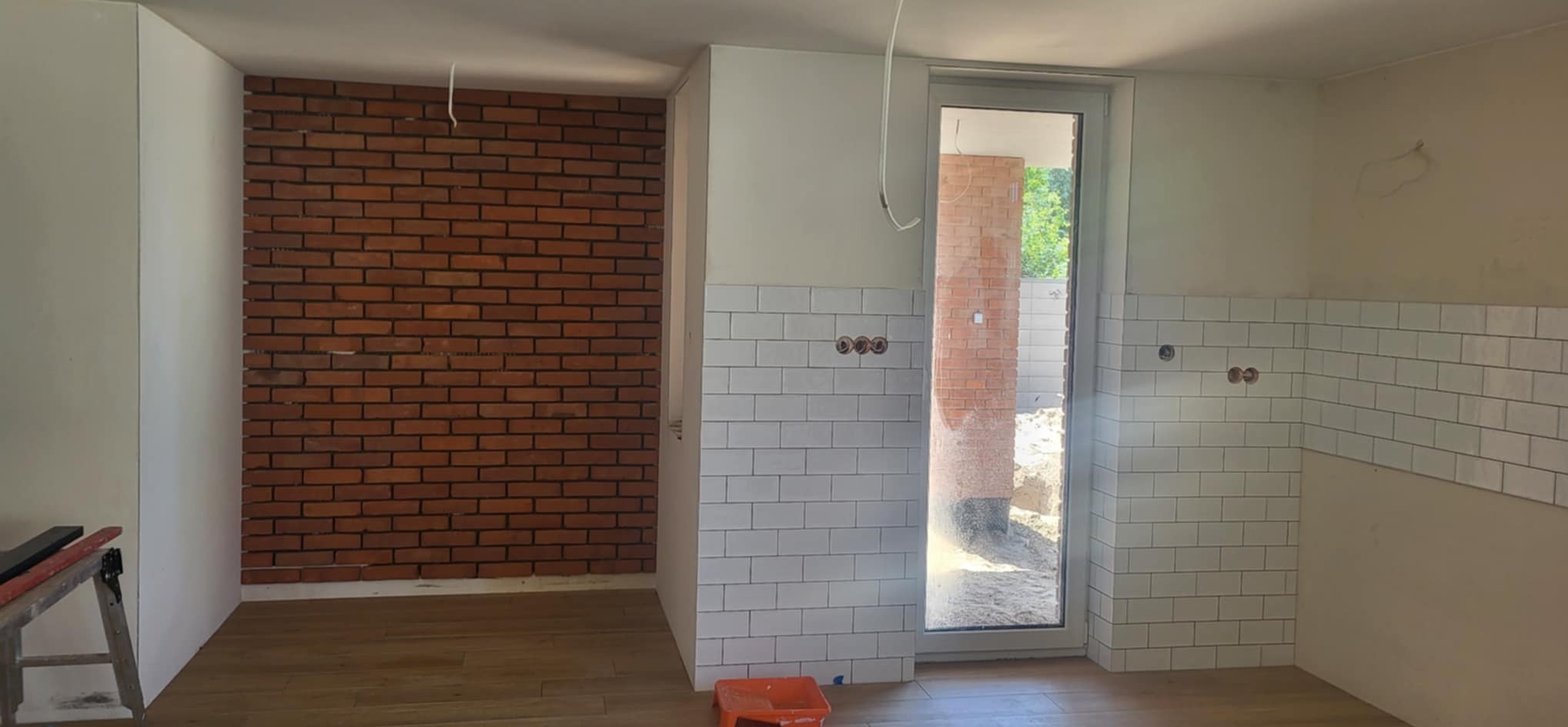 Wnętrze w trakcie wykańczania: ceglana ściana, białe płytki typu metro na ścianie, drewniana podłoga i widoczne instalacje elektryczne.
