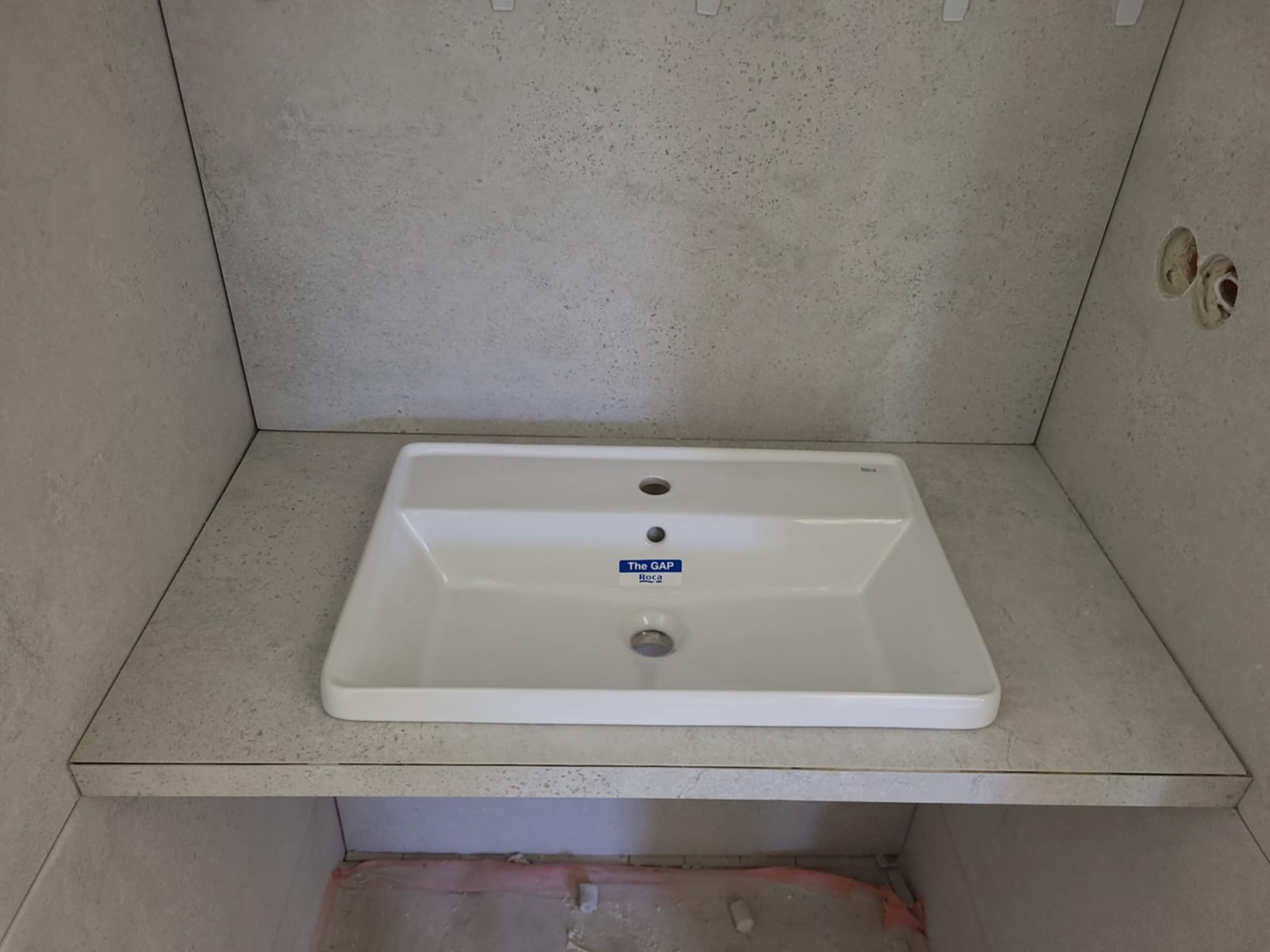 Umywalka Roca The Gap wkomponowana w blat z płytek imitujących kamień, na tle ściany wyłożonej tymi samymi płytkami. Widoczne gniazdka elektryczne.