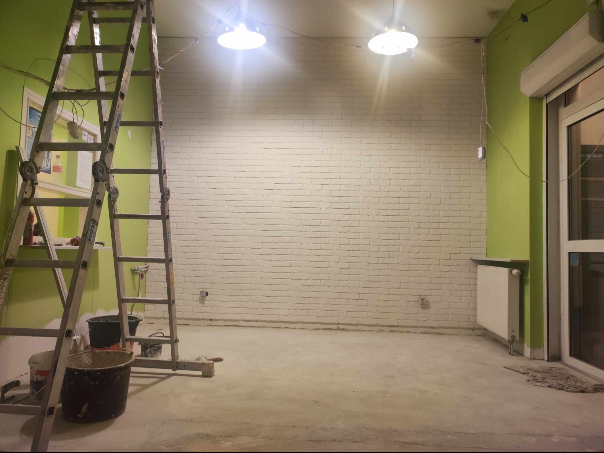 Biała ceglana ściana w trakcie wykańczania, widoczne drabiny, wiadra z farbą i niedomalowane fragmenty na ścianie, surowa podłoga i zielone ściany.