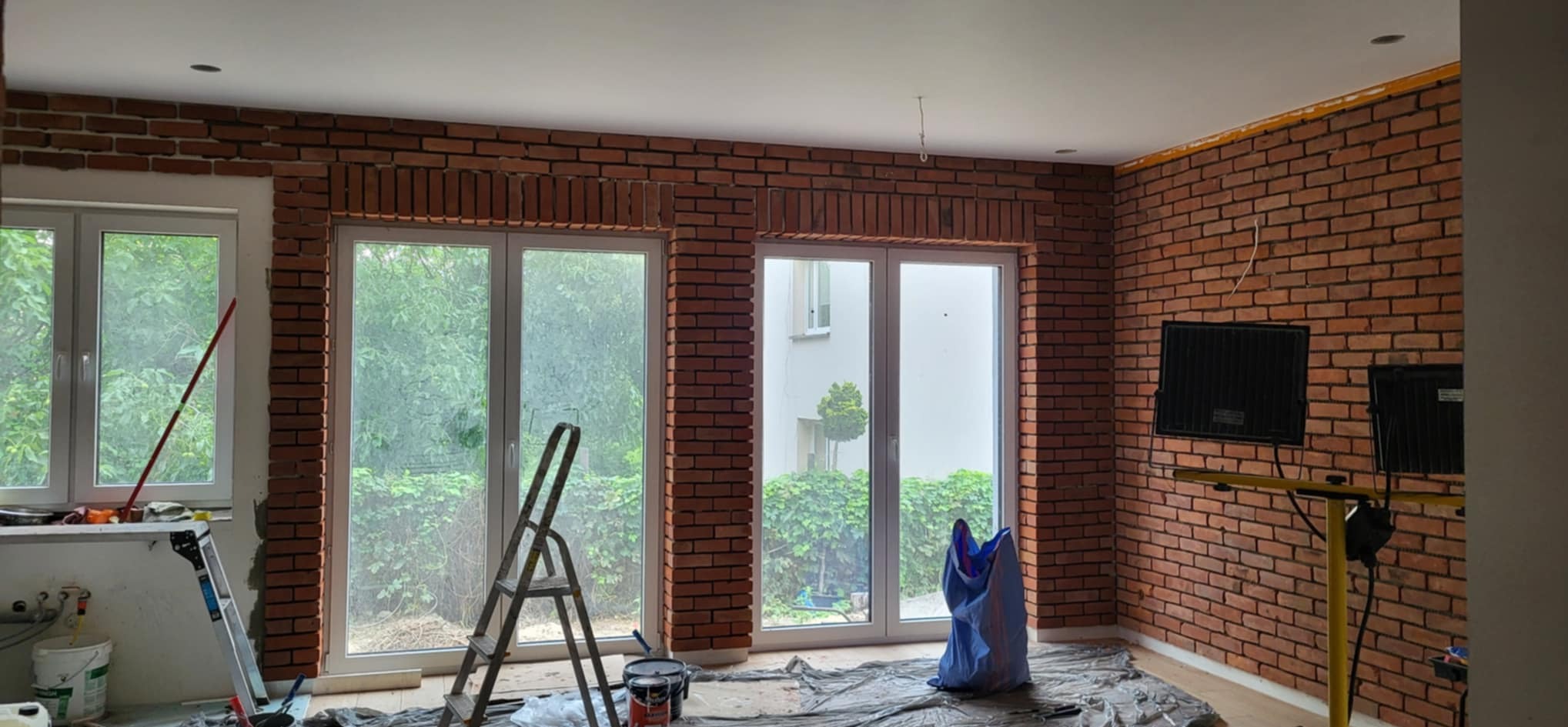 Ściana z cegły klinkierowej w trakcie montażu w jasnym wnętrzu. Widoczne okna, drabina, narzędzia i folia ochronna. Oświetlenie budowlane.