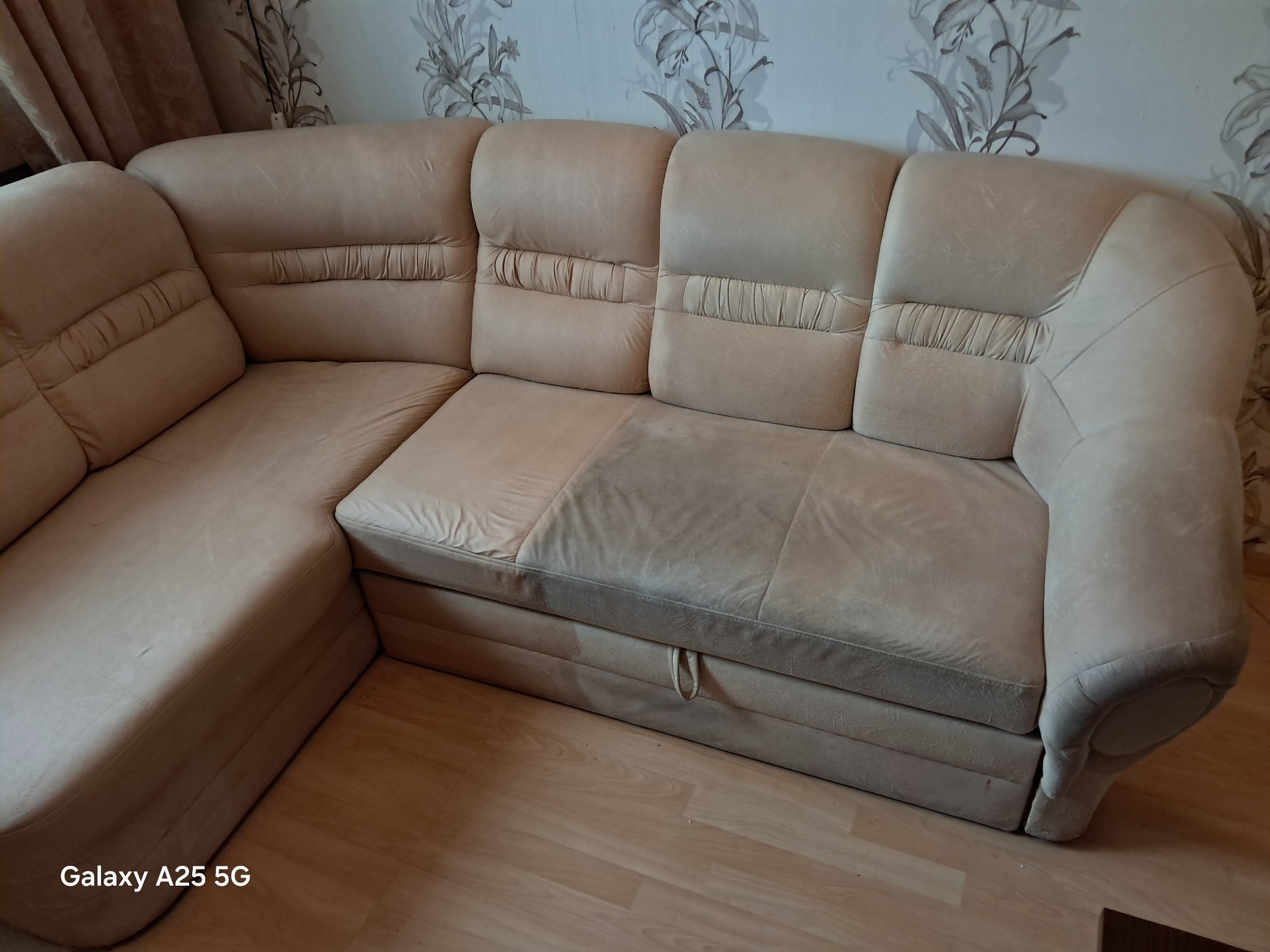 Beżowa, narożna kanapa z widocznym fragmentem po czyszczeniu, ukazującym różnicę w odcieniu. Tapeta w kwiaty w tle. Widoczny fragment podłogi.