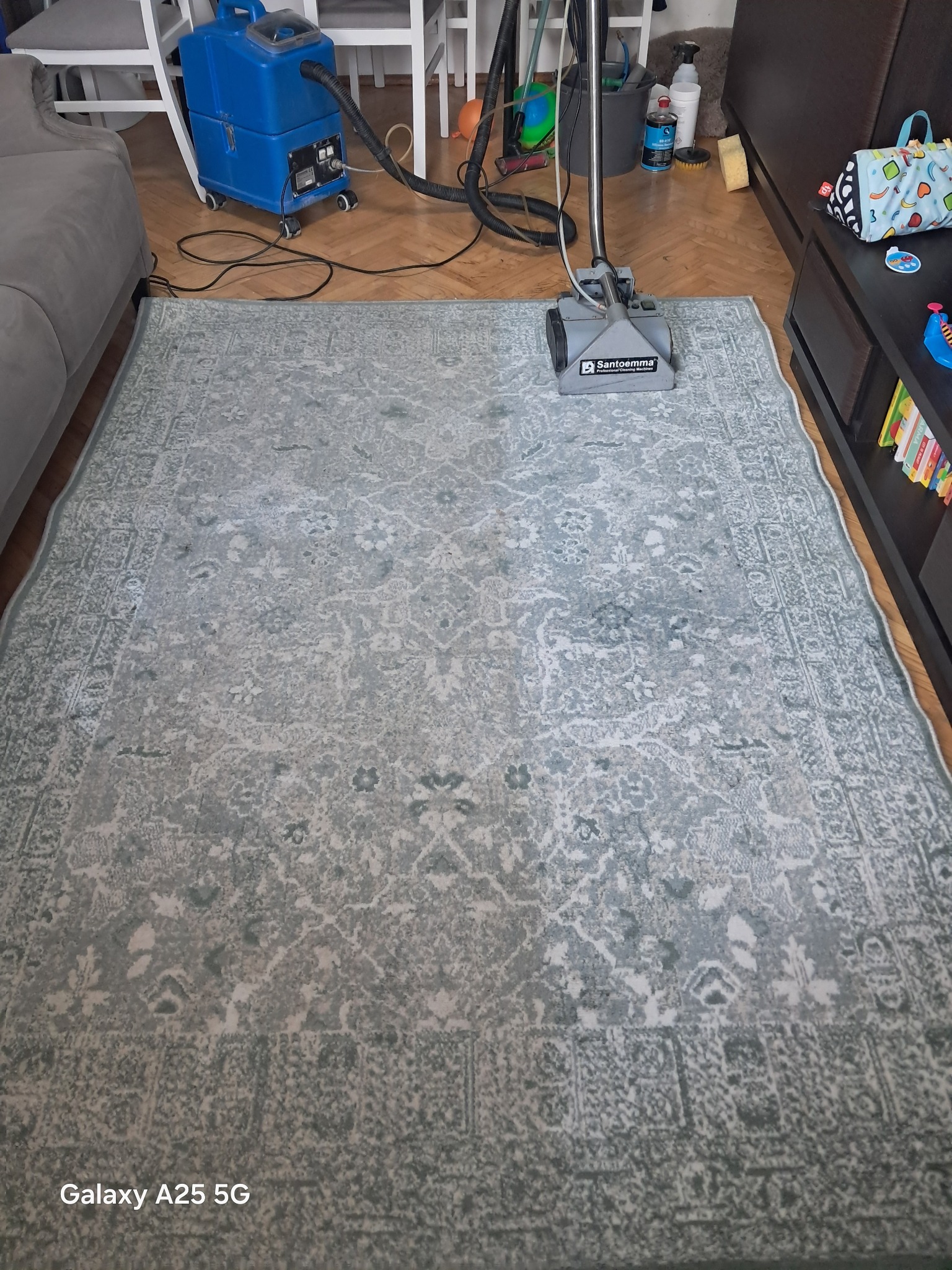 Szary dywan z wzorem czyszczony profesjonalnym odkurzaczem Santoemma. Widoczne meble w tle. Parkiet na podłodze. Sprzęt do czyszczenia dywanów.