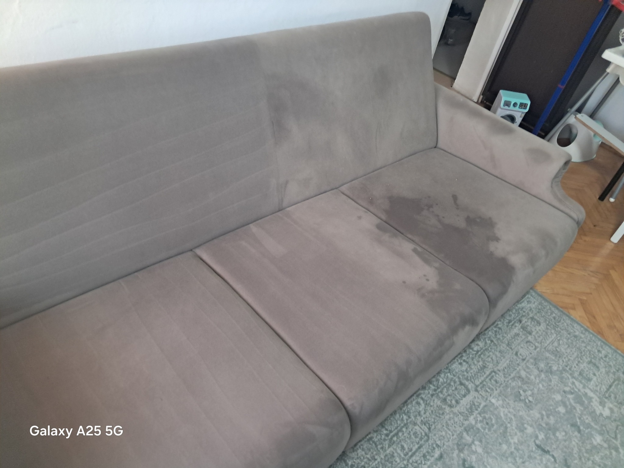 Szara kanapa z widoczną różnicą w czystości tapicerki po lewej stronie, po prawej widoczne plamy. Dywan z geometrycznym wzorem w tle.