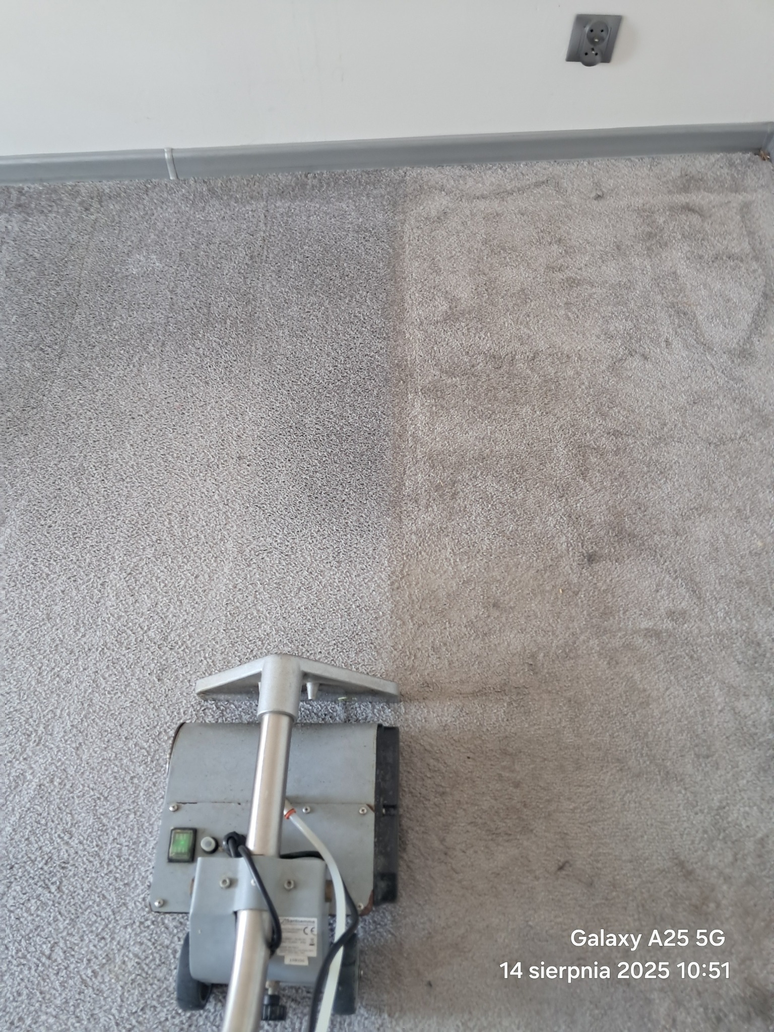 Szary dywan częściowo wyczyszczony, widoczna różnica w odcieniu. Na dole kadru fragment maszyny czyszczącej. U góry listwa przypodłogowa i gniazdko.