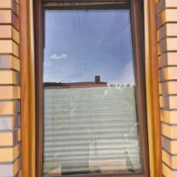 Okno z brązową ramą i plisami. Widoczny fragment muru z cegły. Przez okno widać błękitne niebo i fragment budynku z czerwonym dachem.