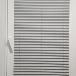 LUXORA - Szare plisy okienne zamontowane w białym oknie, widoczne z bliska. Na parapecie leżą narzędzia i dokumenty, co sugeruje świeży montaż.