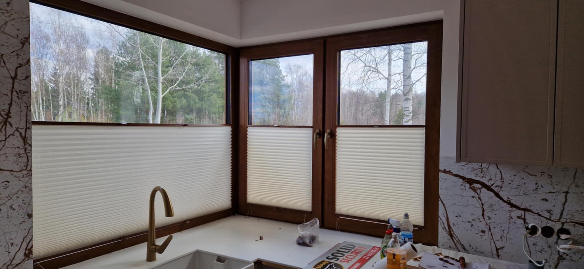 Jasne plisy okienne w brązowych ramach, widok na las za oknem. Złota bateria kuchenna i marmurowy wzór na ścianie dodają elegancji.