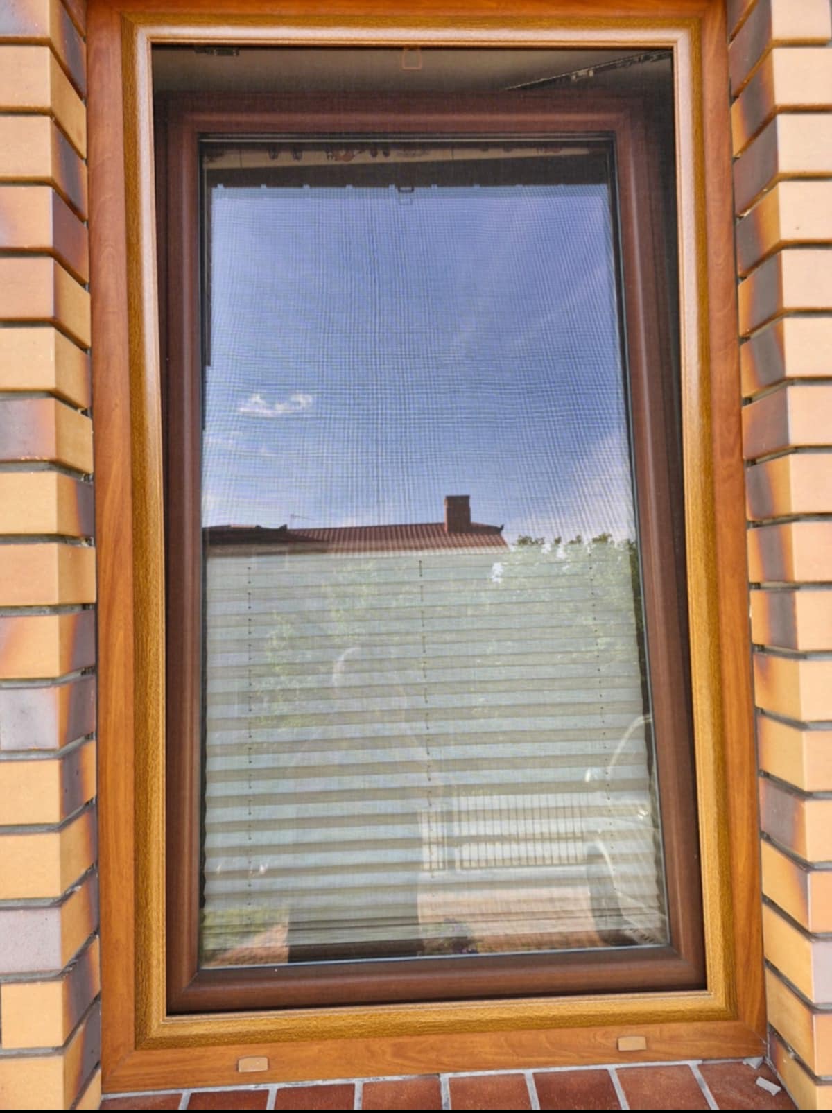 Okno z brązową ramą i plisami. Widoczny fragment muru z cegły. Przez okno widać błękitne niebo i fragment budynku z czerwonym dachem.
