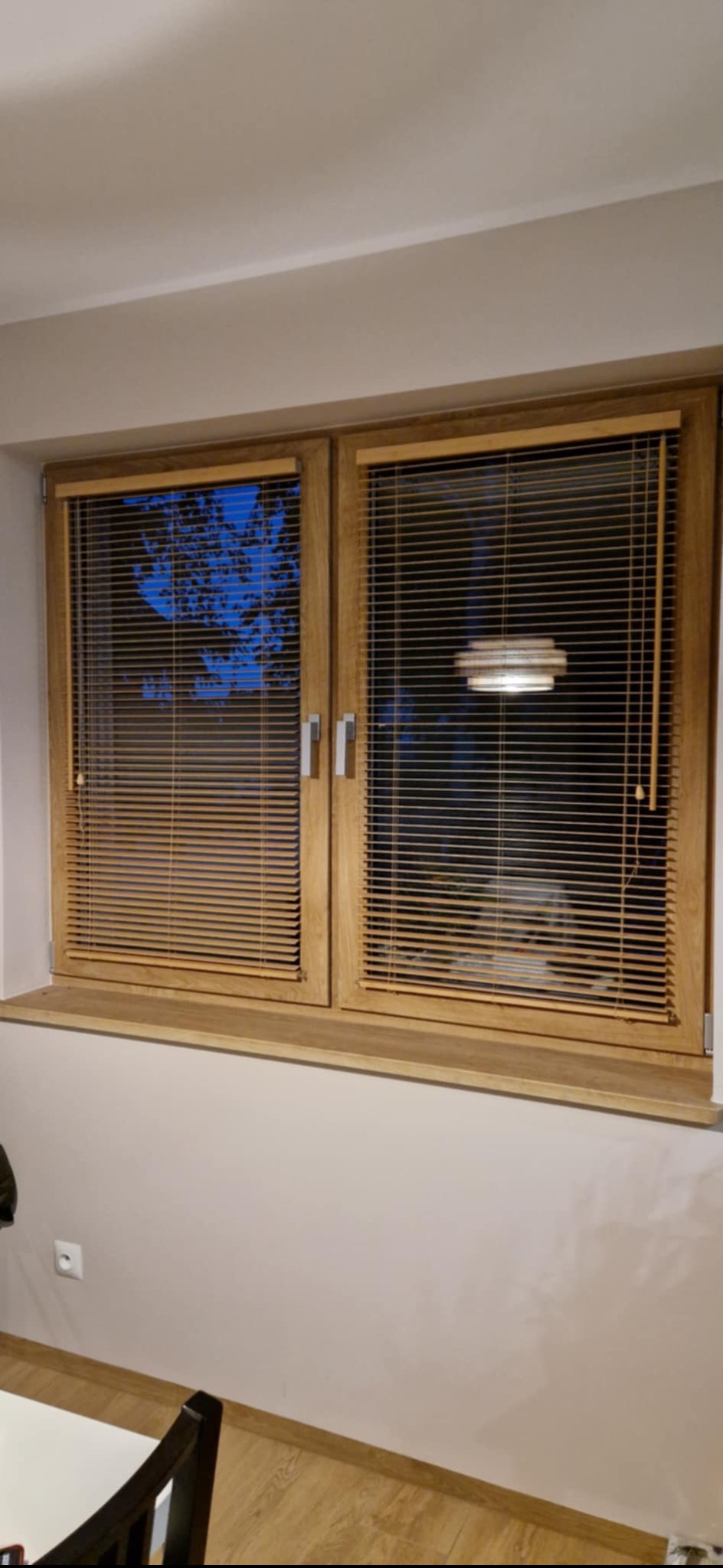 Dwa okna z drewnianymi żaluzjami aluminiowymi, widok na zewnątrz i lampę. Naturalne światło podkreśla strukturę drewna. Detal wnętrza z bliska.