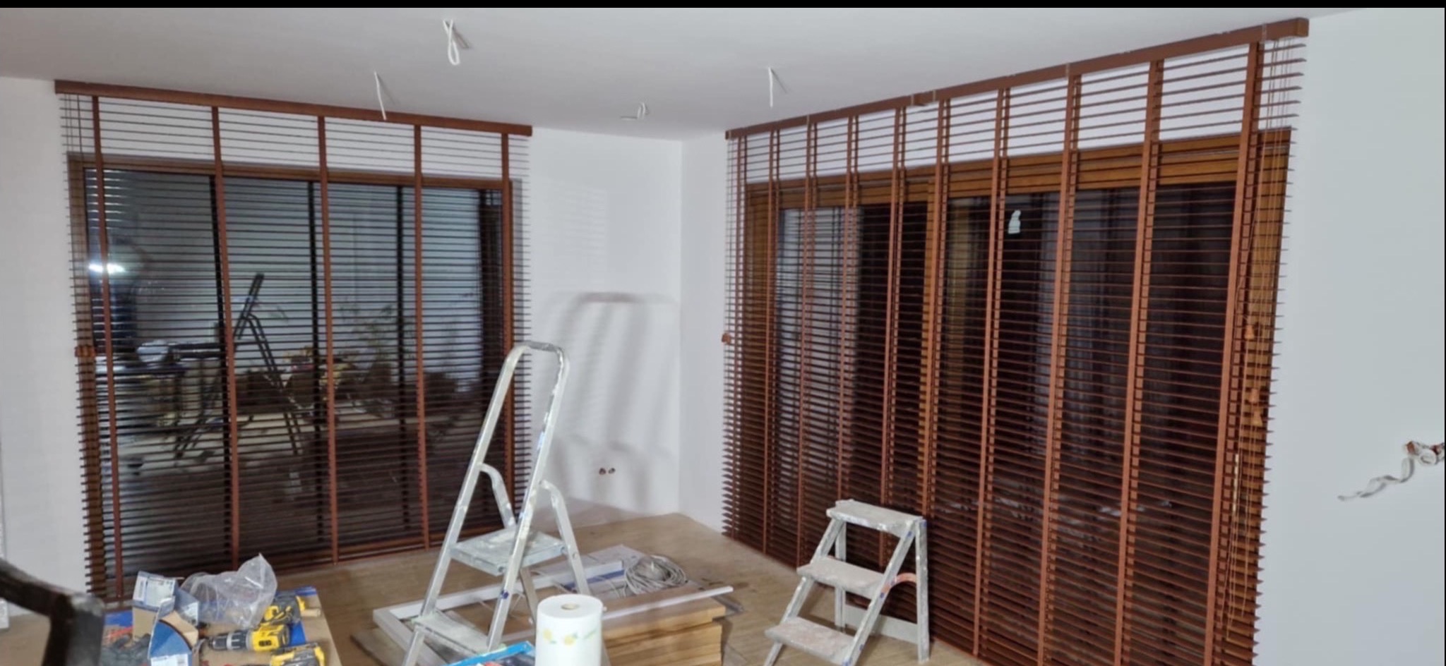 Montaż drewnianych żaluzji w oknach. Wnętrze z drabinami i narzędziami, gotowe osłony okienne z brązowego drewna w jasnym pokoju.