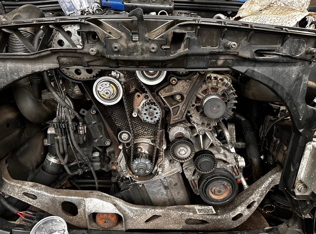 Odsłonięty silnik samochodowy z widocznymi elementami układu rozrządu i osprzętu, ukazujący skomplikowaną budowę i detale mechaniczne jednostki napędowej.