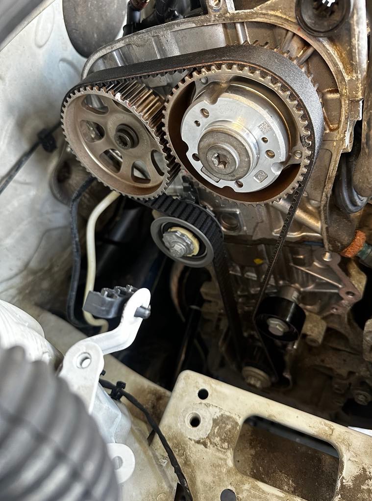 Silnik samochodowy z widocznym paskiem rozrządu i kołami zębatymi. Detal mechanizmu napędowego w trakcie naprawy lub serwisu. Widoczne przewody i elementy mocujące.