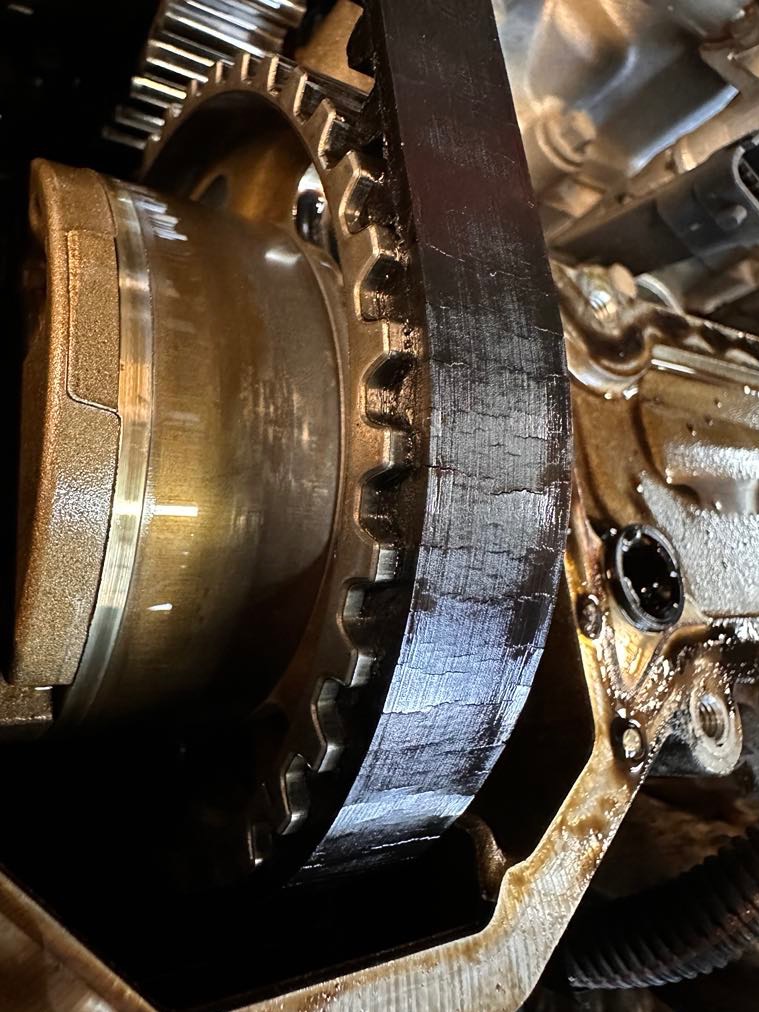 Zużyty pasek rozrządu z widocznymi pęknięciami, założony na zębatym kole w silniku samochodowym. Widoczne ślady oleju.