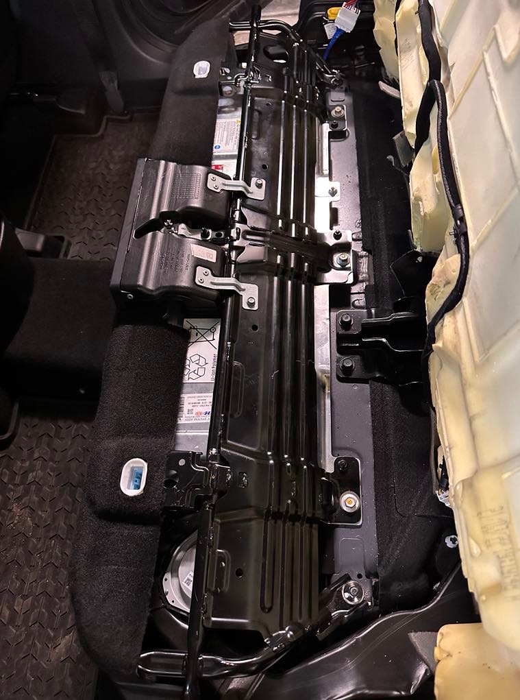 Demontaż tylnej kanapy samochodowej, odsłonięta metalowa konstrukcja z widocznymi śrubami i elementami tapicerki. Widoczny głośnik w podłodze.