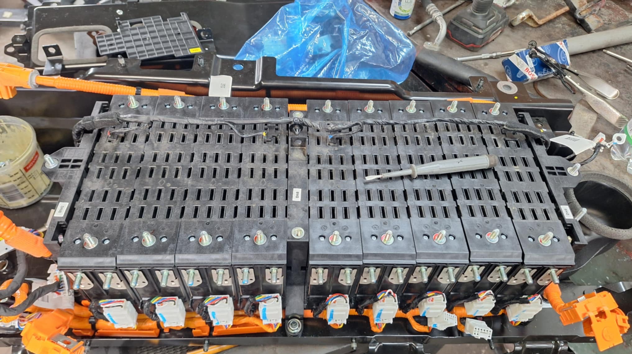 Otwarta bateria samochodowa z pomarańczowymi kablami i śrubokrętem na wierzchu, w warsztacie samochodowym podczas naprawy lub diagnostyki.