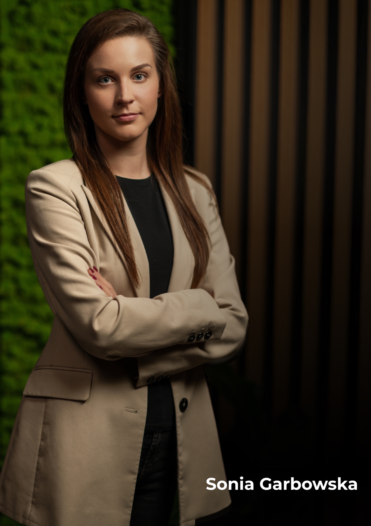 Profesjonalny portret kobiety w beżowym żakiecie na tle ściany z mchu i drewnianych paneli. Skrzyżowane ramiona, pewny wyraz twarzy, oświetlenie studyjne.