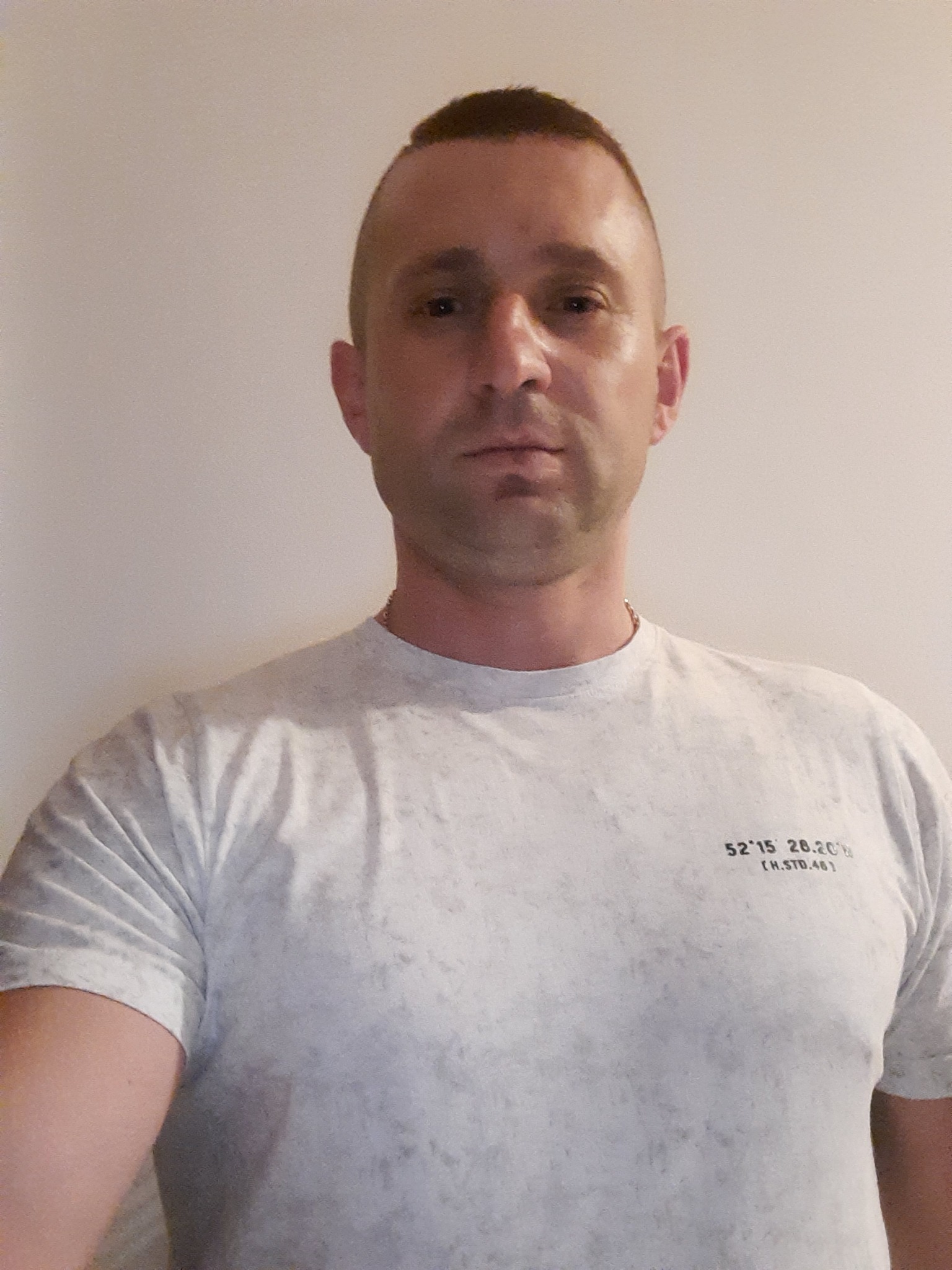 Portret mężczyzny o krótkich włosach w szarej koszulce z nadrukiem współrzędnych na jasnym tle, z delikatnym łańcuszkiem na szyi. Ujęcie od klatki piersiowej.